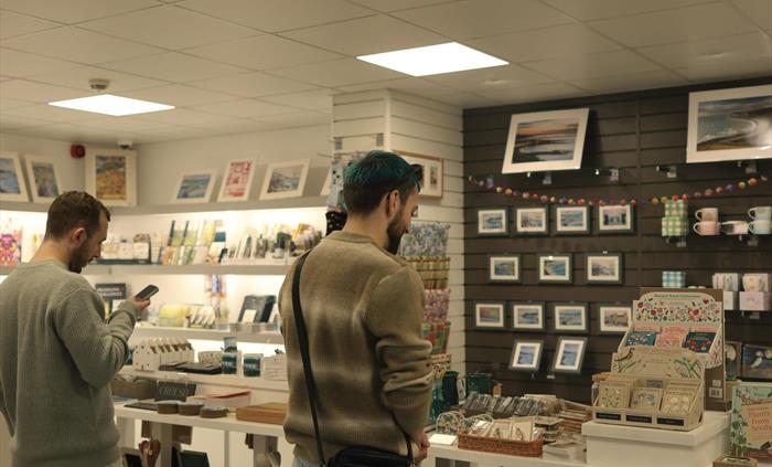 Two men are standing in the shop with a table of a variety of shop items and a wall display in front of them.
